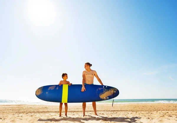 Pai e filho carregam prancha de surf na praia de areia — Fotografia de Stock