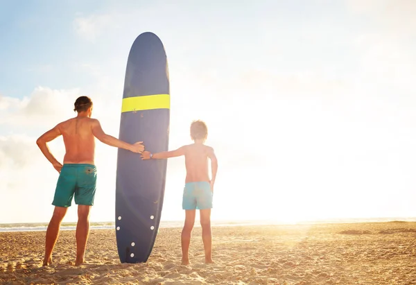 Father and son stand with surf board over sunset — Stockfoto