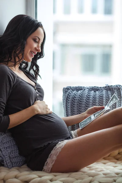Vista lateral de la joven embarazada feliz mirando ultrasonido en alféizar de la ventana —  Fotos de Stock