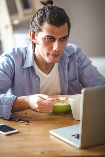 Joven comiendo hojuelas de maíz con leche y mirando a la pantalla del portátil — Foto de Stock