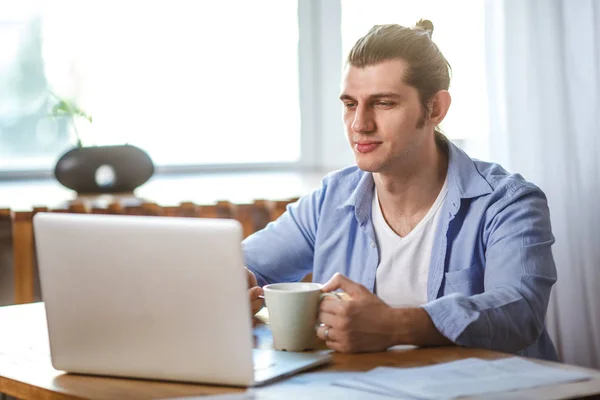Νεαρός άνδρας κρατά την κούπα, κάθεται στο τραπέζι και ψάχνει για την οθόνη του φορητού υπολογιστή — Φωτογραφία Αρχείου