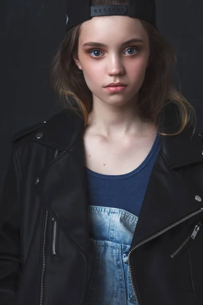 Jovem adolescente usando jeans jaqueta de couro e boné, retrato de estúdio sobre fundo escuro — Fotografia de Stock