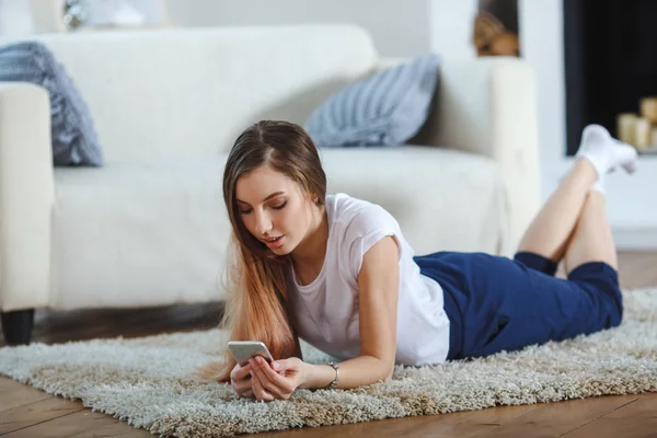 Mujer joven revisando su teléfono inteligente tumbado en la alfombra —  Fotos de Stock