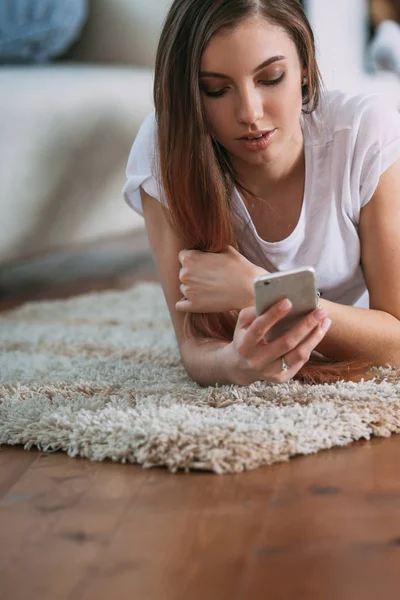 Mujer joven revisando su teléfono inteligente tumbado en la alfombra —  Fotos de Stock