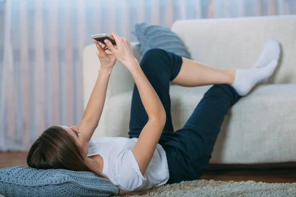 Mujer joven revisando su teléfono inteligente tumbado en la alfombra —  Fotos de Stock