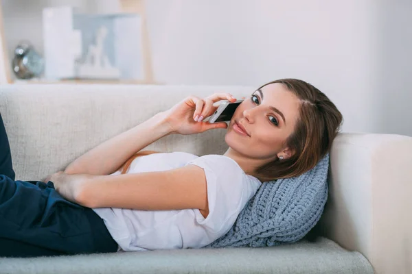 Jovem mulher chamando por seu telefone inteligente deitado no sofá — Fotografia de Stock