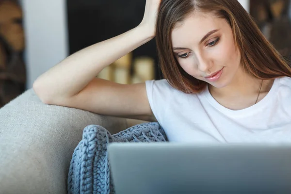 Ung kvinna som sitter med laptop på soffan i sitt hem Stockbild