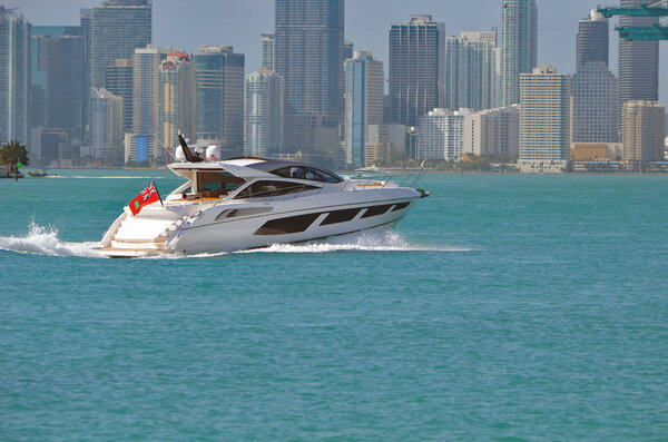 Гладкая моторная яхта направляется к порту Майами с высотными жилыми и коммерческими зданиями на заднем плане.
