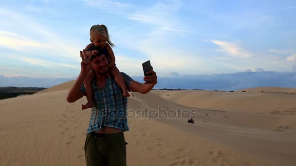 Otec nese malé děvče na Duně closeup dělá Selfie při západu slunce — Stock video