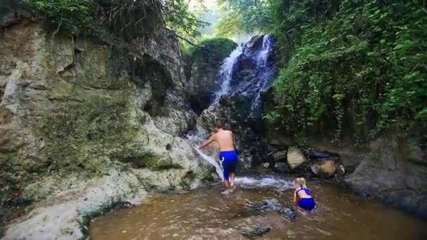 Man klimt tussen rotsen naar waterval en meisje speelt in Water — Stockvideo