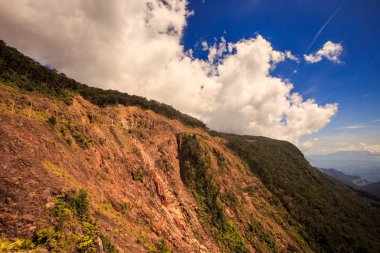 closeup kahverengi taşlı dağ yamacı üstte mavi gökyüzünde beyaz bulutlar karşı tropikal orman