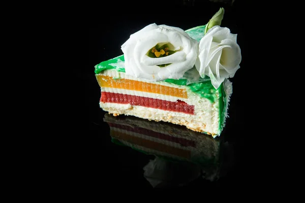 黄色と赤と緑釉のおいしいムース ケーキの上面層間をゼリーし ブラック ミラーの背景を提供しています上の白い花で飾られました — ストック写真