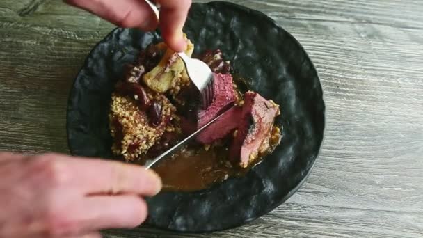 Menschenhände schneiden halb rohes Fleisch mit Gabel und Messer auf schwarzem Teller — Stockvideo