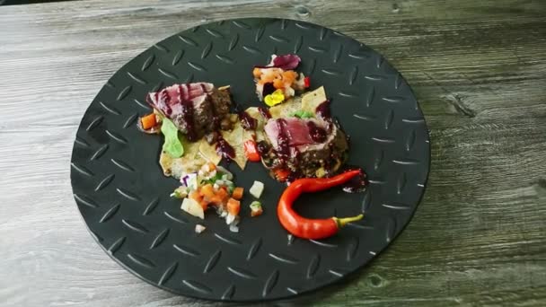 赤唐辛子とソースで飾られたロースト肉の作品でズームは 黒のテクスチャレストランプレート上のポテトチップスや野菜サラダを提供 — ストック動画