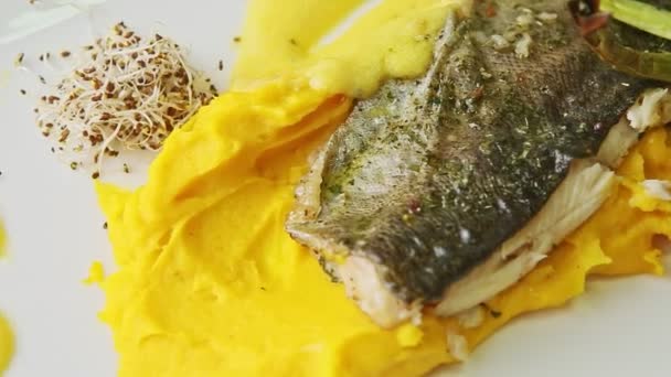 Крупный план жареного филе морской рыбы на картофельном пурпуре и нарезанных овощей вращается на тарелке — стоковое видео