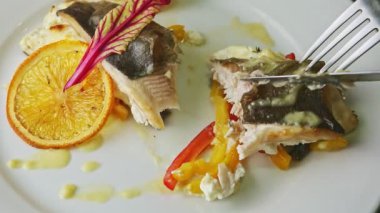 closeup hands cut off small piece from baked sea fish by fork and knife on plate