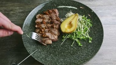 human hands cut off one slice of grilled meat by fork and knife on black plate