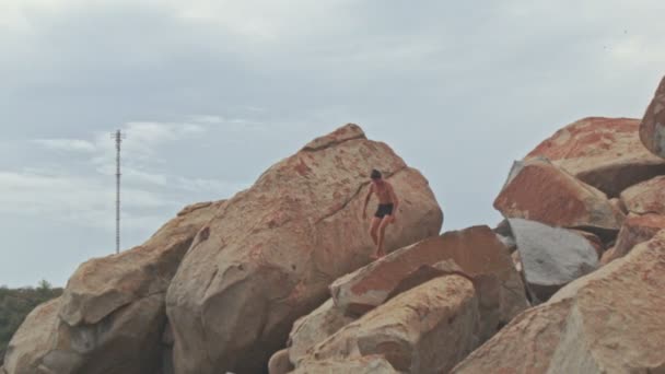 Парень стоит на большом высоком камне на пляже у подножия холма — стоковое видео