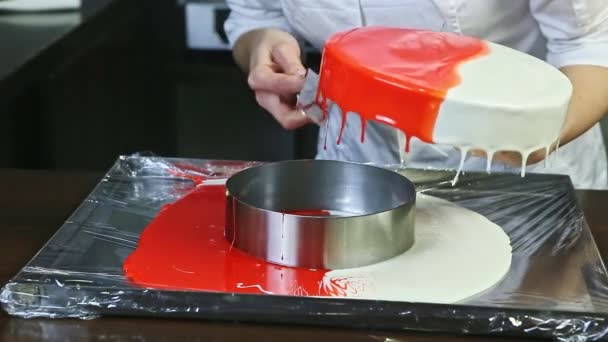 シェフの制服を着たお菓子は キッチンテーブルの上の金属製スタンドに赤と白のガラス製の丸いチーズケーキから長いナイフで余分な釉薬を除去します — ストック動画