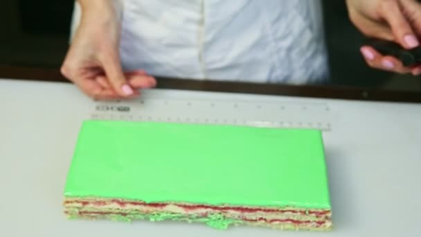 Confectioner in chef uniform puts ruler next to green glazed rectangular layered cake — Stockvideo