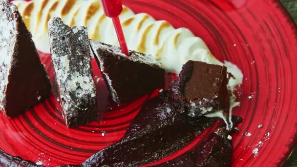 Primer plano mano humana toma por tenedor pedazo de pastel de brownie con crema de plato rojo — Vídeo de stock