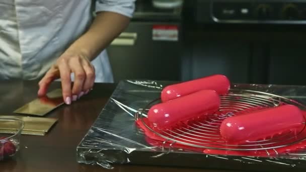 Banketbakker door handen met mes zet roze geglazuurd ovaal dessert op gouden standaard — Stockvideo