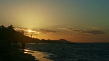 Sarı güneş büyük bulutların ve palmiye ağaçlarının arkasında batıyor deniz kıyısında.