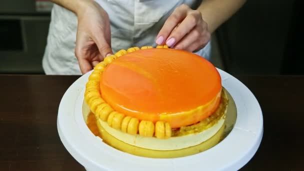 Кондитер руками украшает оранжевый глазированный чизкейк маленькими макаронами — стоковое видео