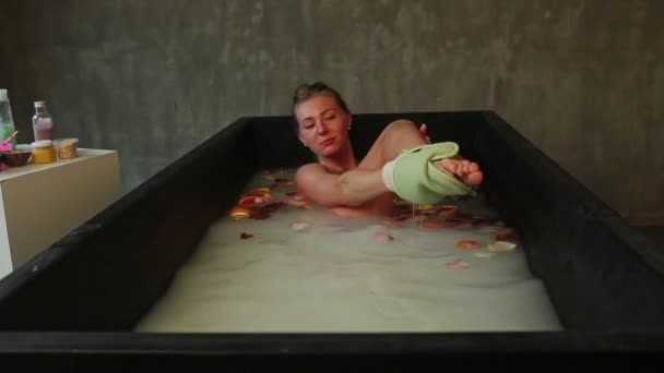 Молодая красивая блондинка моет ногу принимая ванну с молоком и лепестками роз — стоковое видео