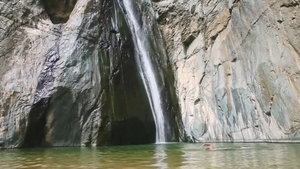 Långsamt panorama upp från ung man simmar i transparent vatten till vitt vattenfall — Stockvideo