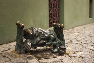 Wroclaw, Polonya - 31 Ekim 2019. Minyatür heykel, sokakta bir yatakta uyuyan cüce. Eski Wroclaw şehrinin merkezinde bulunan bronz cüce, yeşil duvar arka planı.
