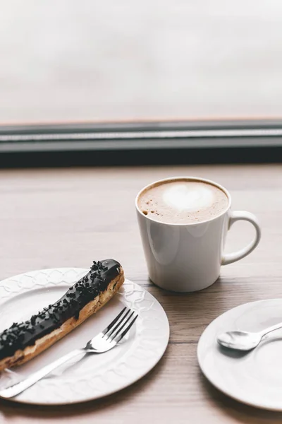 Taza de café sobre mesa de madera con eclair — Foto de Stock