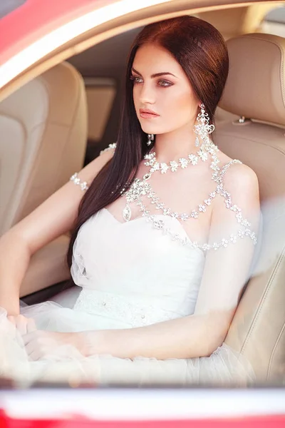 Νεαρή μελαχρινή νύφη σε λευκό φόρεμα κάθεται και θέτοντας σε ένα αυτοκίνητο, ένα όμορφα αξεσουάρ — Φωτογραφία Αρχείου
