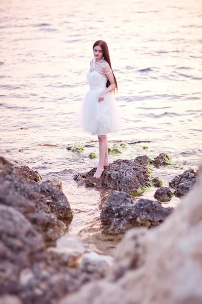 Jeune mariée brune en robe blanche posant au bord de la mer au coucher du soleil — Photo