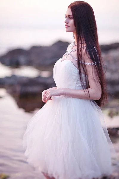 Νέοι μελαχρινή νύφη με λευκό φόρεμα ποζάρει δίπλα στη θάλασσα στο ηλιοβασίλεμα — Φωτογραφία Αρχείου
