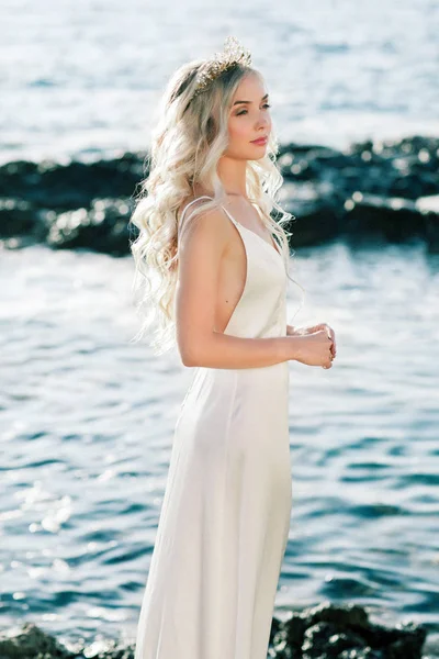 Lovely blond bride in white silk wedding dress posing near the sea — Stock Photo, Image