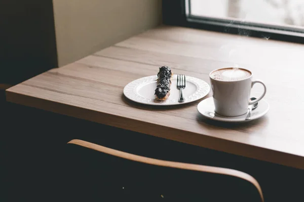 Taza de café sobre mesa de madera con eclair — Foto de Stock