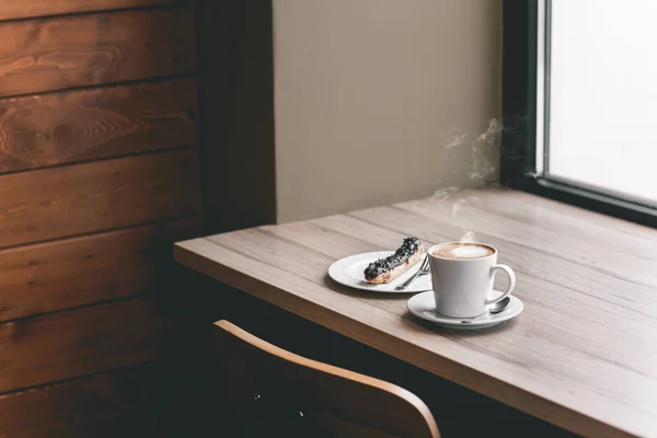 Taza de café sobre mesa de madera con eclair —  Fotos de Stock