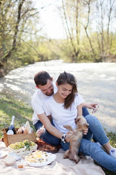 Pique-nique sur l'herbe près de la rivière le jour d'été paresseux. Jeune couple souriant avec petit chien relaxant sur la nature — Photo