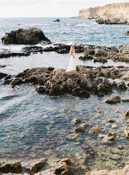 Blond Panna Młoda pozowanie na skały w pobliżu morza. Delikatny i szczere zdjęcia. Ślub za granicą. Ślub na plaży — Zdjęcie stockowe
