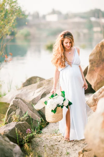 Vackra gravid flicka med långt hår gå nära stranden av en sjö med blommor i en sommarklänning — Stockfoto