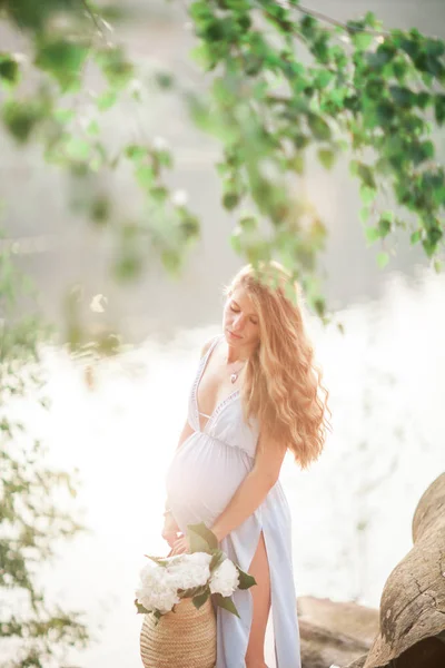 Bela menina grávida com cabelos longos andando perto da costa de um lago com flores em um vestido de verão — Fotografia de Stock