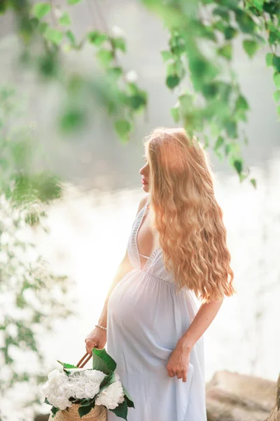 Bela menina grávida com cabelos longos andando perto da costa de um lago com flores em um vestido de verão — Fotografia de Stock