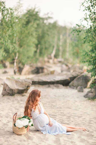 Krásná těhotná dívka s dlouhými vlasy, chůze v blízkosti břehu jezera s květinami v letních šatech — Stock fotografie