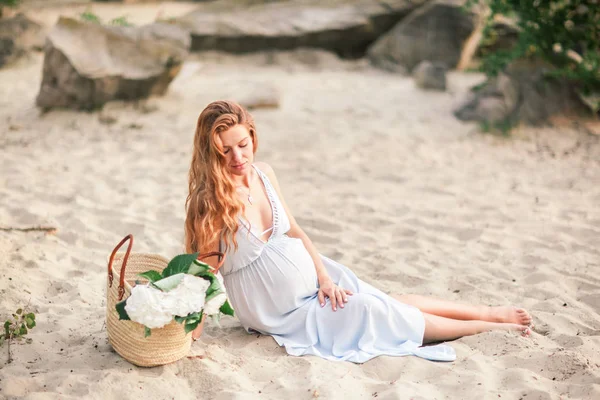 Bela menina grávida com cabelos longos andando perto da costa de um lago com flores em um vestido de verão — Fotografia de Stock