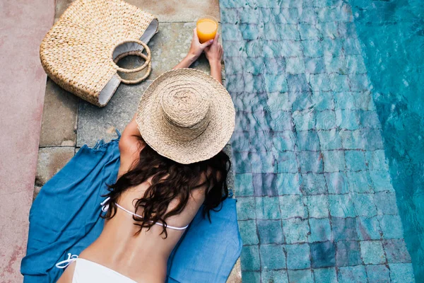Γυναίκα σε ψάθινο καπέλο χαλαρώνοντας στην πισίνα με χυμό πορτοκαλιού. — Φωτογραφία Αρχείου