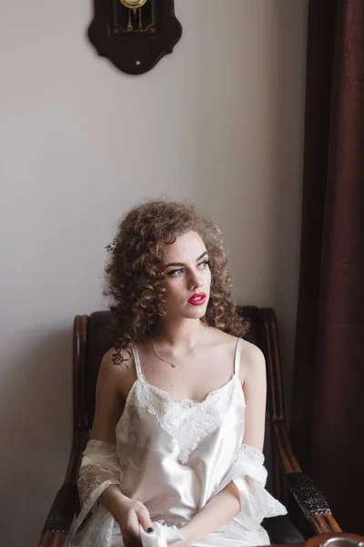 The curly-haired girl sits on an armchair at it the class figure and she is dressed in a dressing-gown — Stock Photo, Image