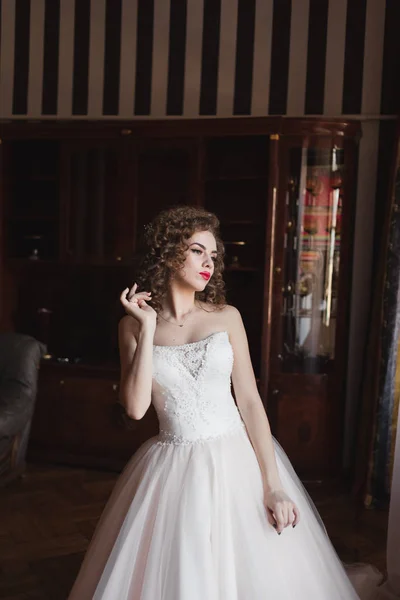 Елегантна дівчина стоїть біля вікна, одягнена у весільну сукню вона має кучеряве волосся — стокове фото