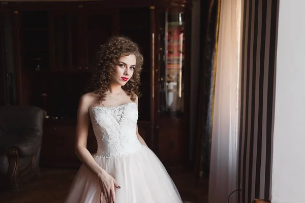 Елегантна дівчина стоїть біля вікна, одягнена у весільну сукню вона має кучеряве волосся — стокове фото