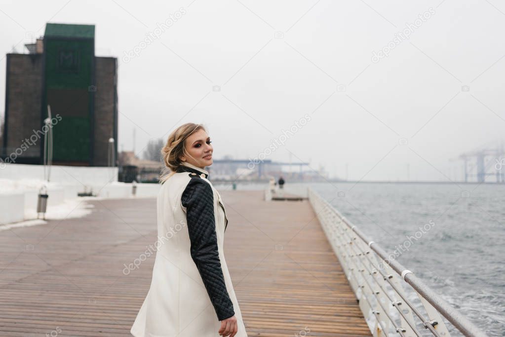 Young blonde girl is standing on the waterfront and is smiling cute.